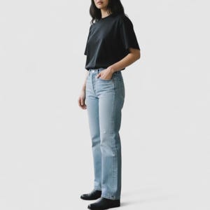 May include: A person wearing a black crew neck t-shirt and light blue denim jeans. The jeans have a straight leg fit. The person is also wearing black leather boots. The background is plain white.