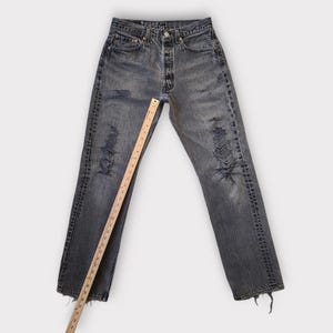 May include: Faded black denim jeans with distressed details, featuring ripped sections and a button fly. A wooden ruler, marked in inches, is placed across the jeans to measure the length. The jeans have a classic five-pocket design.