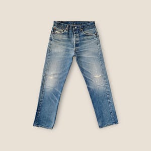 Könnte beinhalten: Eine blaue Jeans aus Denim mit hoher Taille und Knopfleiste. Der Stoff zeigt eine verwaschene Optik mit helleren Stellen an den Hüften sowie dezente Abnutzungserscheinungen, einschließlich kleiner ausgefranster Bereiche an Knien und Taschen.