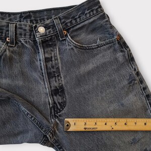 May include: Close-up of a pair of faded black denim jeans. The jeans have a button closure, pockets, and distressed details. A wooden ruler shows measurements in inches.
