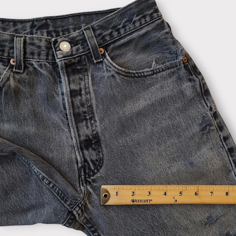 May include: Close-up of a pair of faded black denim jeans. The jeans have a button closure, pockets, and distressed details. A wooden ruler shows measurements in inches.