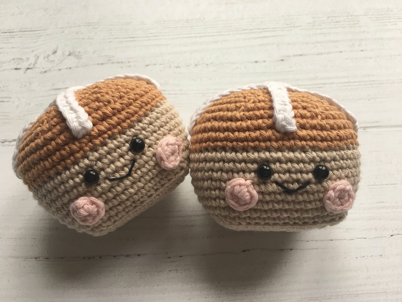 May include: Two crocheted hot cross buns with smiling faces. The buns are brown and beige with white icing and pink cheeks.