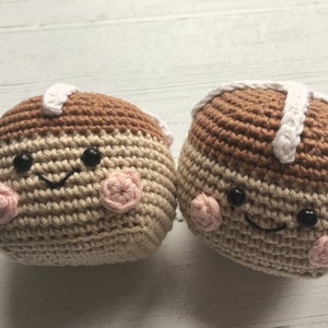 May include: Two crocheted plush toys shaped like hot cross buns with smiling faces. The buns are brown and beige with white icing and pink cheeks.
