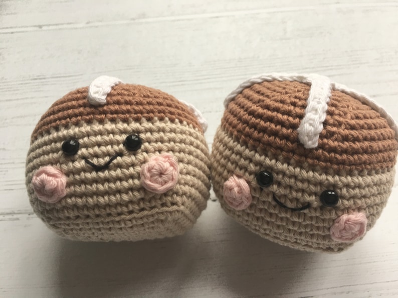 May include: Two crocheted plush toys shaped like hot cross buns with smiling faces. The buns are brown and beige with white icing and pink cheeks.