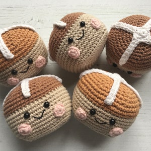 May include: Five crocheted hot cross bun plushies with smiling faces. The plushies are brown and beige with white cross details.