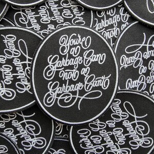 May include: A close-up of several black embroidered patches with white stitching. The patches are round and feature the text "You're a Garbage Can, not a Garbage Canto".