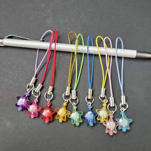 May include: A collection of colorful star-shaped charms with silver-tone metal clasps and matching cords. The charms are in various colors, including purple, red, yellow, green, blue, pink, and light blue. A silver mechanical pencil rests on top.