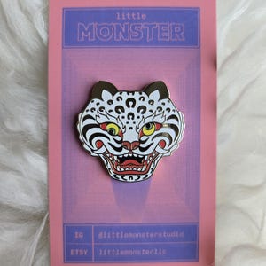 May include: A detailed enamel pin featuring a stylized tiger head with white, black, red, and yellow accents. The pin is set against a pink card with the text "little MONSTER" and social media handles.