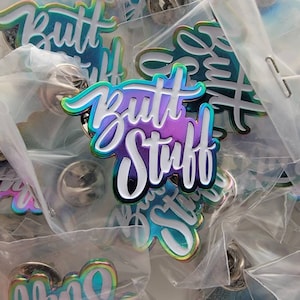 May include: A collection of enamel pins with the words "Butt Stuff" in a cursive font. The pins have a gradient color scheme of purple, blue, and green with a white outline. Each pin is individually packaged in a clear plastic bag.