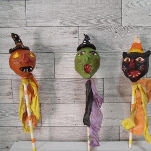 May include: Three Halloween-themed paper mache figures on sticks. The figures include a jack-o'-lantern, a green witch, and a black cat. Each figure wears a hat and has colorful fabric accents. The background is a light wood-grain pattern.