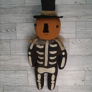 May include: A fabric doll featuring a round orange pumpkin head with a black top hat, a brown nose, and black bead eyes. The body is black with a painted white skeleton rib cage and bone pattern, accented by a tan frayed collar around the neck.