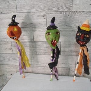 May include: Three Halloween-themed stick figures. The first is an orange pumpkin with a black witch hat. The second is a green witch with a black hat. The third is a black cat with a witch hat. Each figure has a colorful fabric tail.