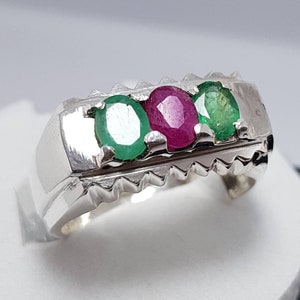 May include: A silver ring with three gemstones. The center stone is a pink ruby, and the two outer stones are green emeralds.