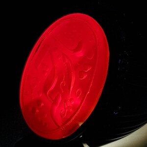 May include: A red oval-shaped wax seal with a decorative design. The seal is made of red wax and has a raised design.