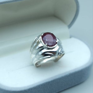 Op de afbeelding: Een zilveren ring met een grote, ovale robijn. De ring heeft een textuur met een golfachtig ontwerp op de band. De robijn is dieprood en is gezet in een eenvoudige bezel setting. De ring wordt gepresenteerd in een grijze sieradendoos.
