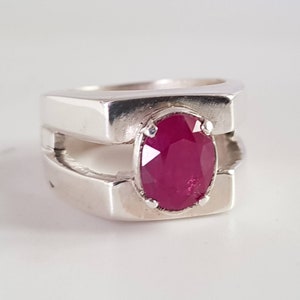 May include: A silver ring with a red oval gemstone set in the center. The ring has a unique design with two bands that intersect at the top and bottom of the stone.
