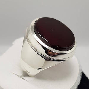 May include: A silver ring with a dark red oval gemstone set in a raised bezel. The ring has a polished finish and a wide band.