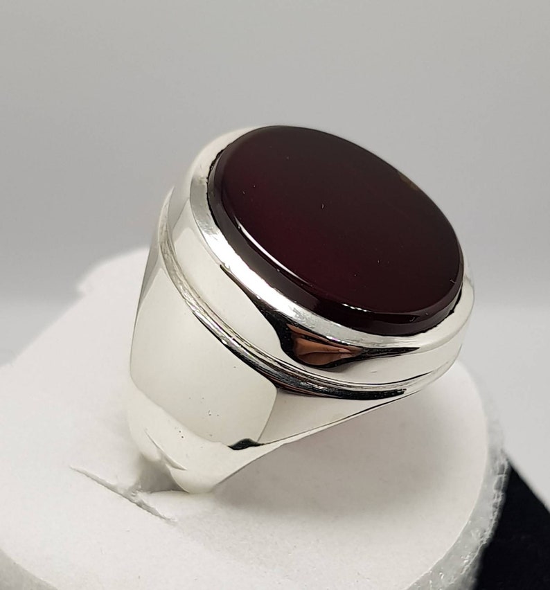 May include: A silver ring with a dark red oval gemstone set in a raised bezel. The ring has a polished finish and a wide band.
