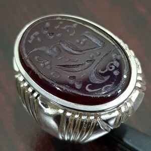 May include: A silver ring with an oval-shaped, dark red gemstone. The gemstone has intricate carvings of Arabic script and floral designs.