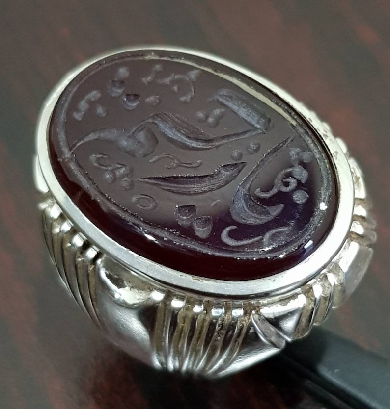 May include: A silver ring with an oval-shaped, dark red gemstone. The gemstone has intricate carvings of Arabic script and floral designs.
