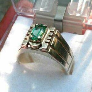 May include: A silver ring with a green emerald gemstone set in a rectangular shape. The ring has a textured band with a stepped design.