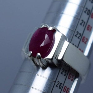 May include: A silver ring with a large, oval-shaped ruby gemstone. The ring is sized on a ring sizer, showing a size of 25.5.