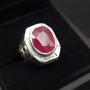 May include: A silver ring with a large, oval-shaped, red ruby gemstone set in the center. The ring has intricate details around the gemstone.