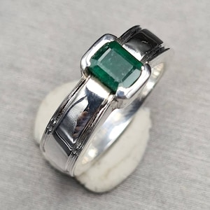 Può includere: Un anello d'argento con uno smeraldo verde incastonato in una montatura quadrata. L'anello ha una fascia testurizzata.