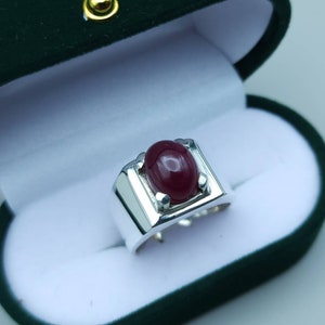 Puede incluir: Un anillo de plata con una gran piedra preciosa de rubí de forma ovalada engastada en un bisel cuadrado. El anillo está en una caja de terciopelo verde.