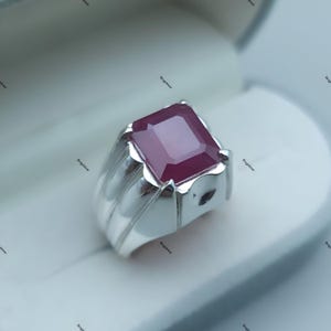 May include: A silver ring with a large, rectangular, deep red gemstone. The ring has a thick band with vertical grooves and a bezel setting that holds the stone securely. The gemstone is the focal point, catching the light.