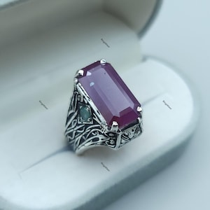 May include: A silver ring featuring a large, rectangular, deep red gemstone. The ring has an ornate, detailed design on the band and a small, green gemstone accent. The ring is presented in a white jewellery box.
