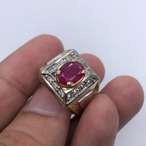 Könnte beinhalten: Ein Silber- und Goldring mit einem großen ovalen Rubin-Edelstein, der von kleineren weißen Edelsteinen umgeben ist.