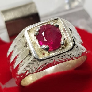May include: A silver ring featuring a red ruby gemstone set in a square-shaped bezel. The ring band has a detailed engraved design.