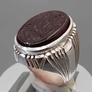 May include: A silver ring with a dark red oval gemstone set in the center. The gemstone has intricate carvings and a silver band with a textured design.