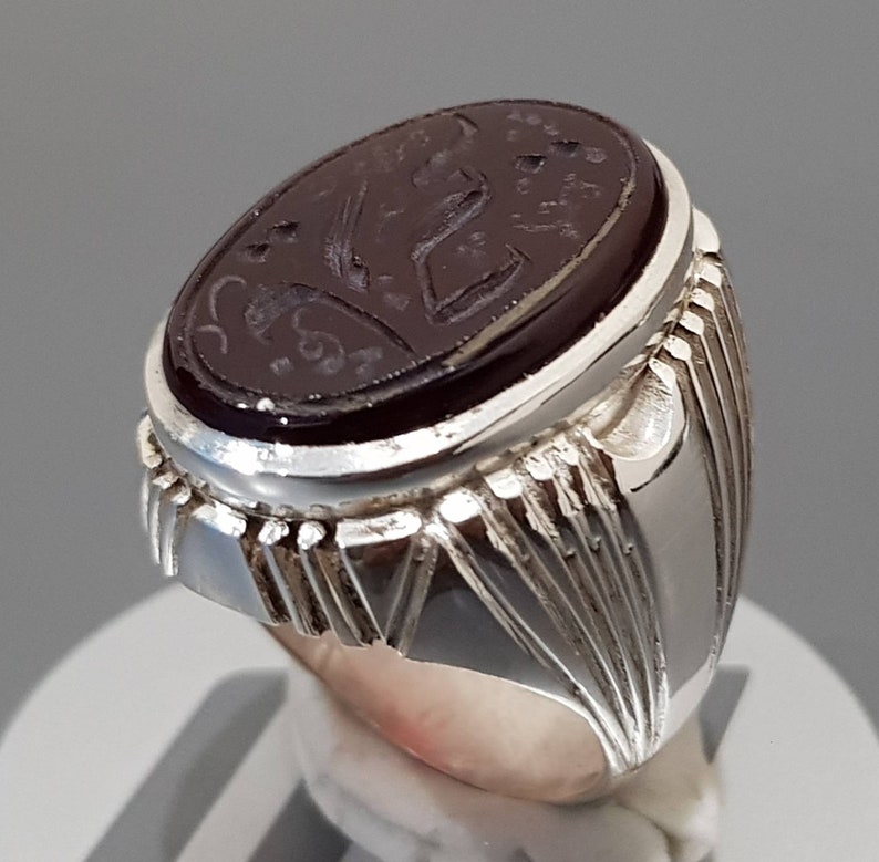 May include: A silver ring with a dark red oval gemstone set in the center. The gemstone has intricate carvings and a silver band with a textured design.
