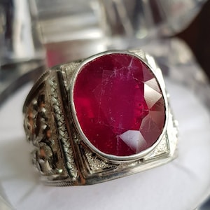 May include: A silver ring with a large, oval-shaped ruby gemstone. The ring has intricate details on the band.