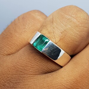 May include: A silver ring with a green emerald gemstone. The ring is on a person's finger.