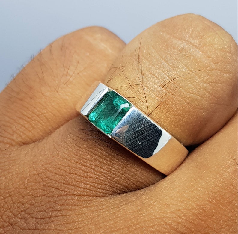May include: A silver ring with a green emerald gemstone. The ring is on a person's finger.