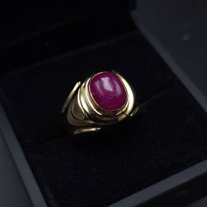 May include: A gold ring with a large, oval-shaped ruby gemstone. The ring has a thick band with a decorative design.