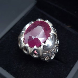 Puede incluir: Un anillo de plata con una gran piedra preciosa de rubí de forma ovalada. El anillo tiene un diseño detallado y adornado.