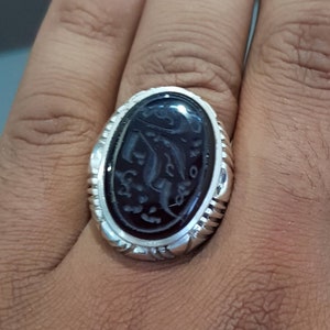 May include: A silver ring with an oval-shaped black onyx stone featuring intricate Arabic calligraphy.