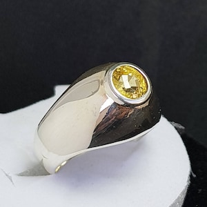 May include: A silver ring featuring a yellow gemstone set in a round bezel. The ring has a wide band and a smooth, polished finish.