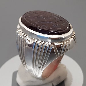 May include: A silver ring with an oval-shaped black stone set in the center. The stone has intricate carvings and is surrounded by a silver band with a decorative design.
