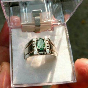 May include: A silver ring with a green oval gemstone set in a silver band. The ring is in a clear plastic box.