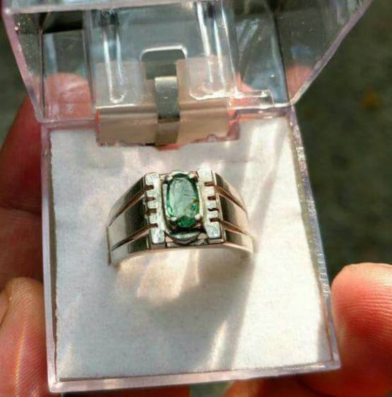 May include: A silver ring with a green oval gemstone set in a silver band. The ring is in a clear plastic box.