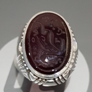 May include: A silver ring with a dark red oval gemstone featuring intricate carved Arabic script.