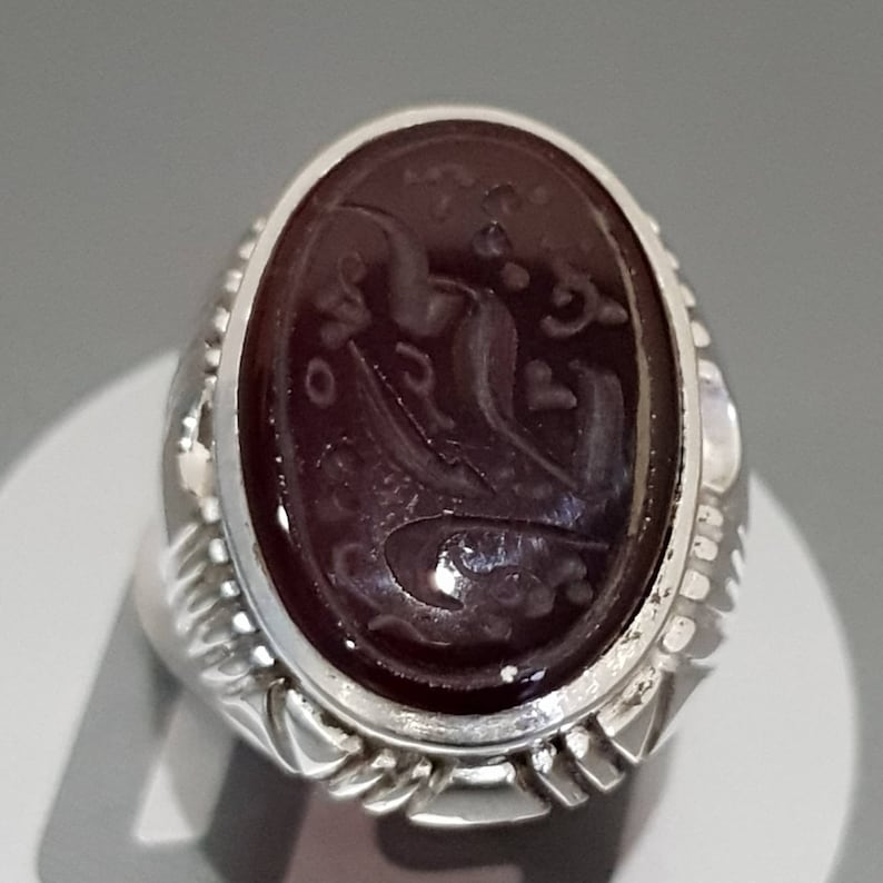 May include: A silver ring with a dark red oval gemstone featuring intricate carved Arabic script.