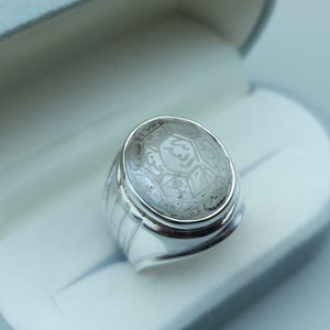 May include: A silver ring with an oval, clear stone. The stone has an intricate, etched design. The ring is set in a silver band with a ribbed design on the sides. The ring is displayed in a white jewelry box.