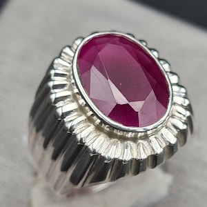 Puede incluir: Un anillo de plata con una gran piedra preciosa de rubí de forma ovalada. El anillo tiene una banda texturizada con un borde decorativo alrededor de la piedra.