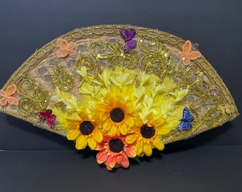 Peut inclure: Un éventail décoratif en matériau tissé avec des accents dorés et orné de papillons et de tournesols. L'éventail est décoré de papillons jaunes, orange, rouge, bleu et violet.